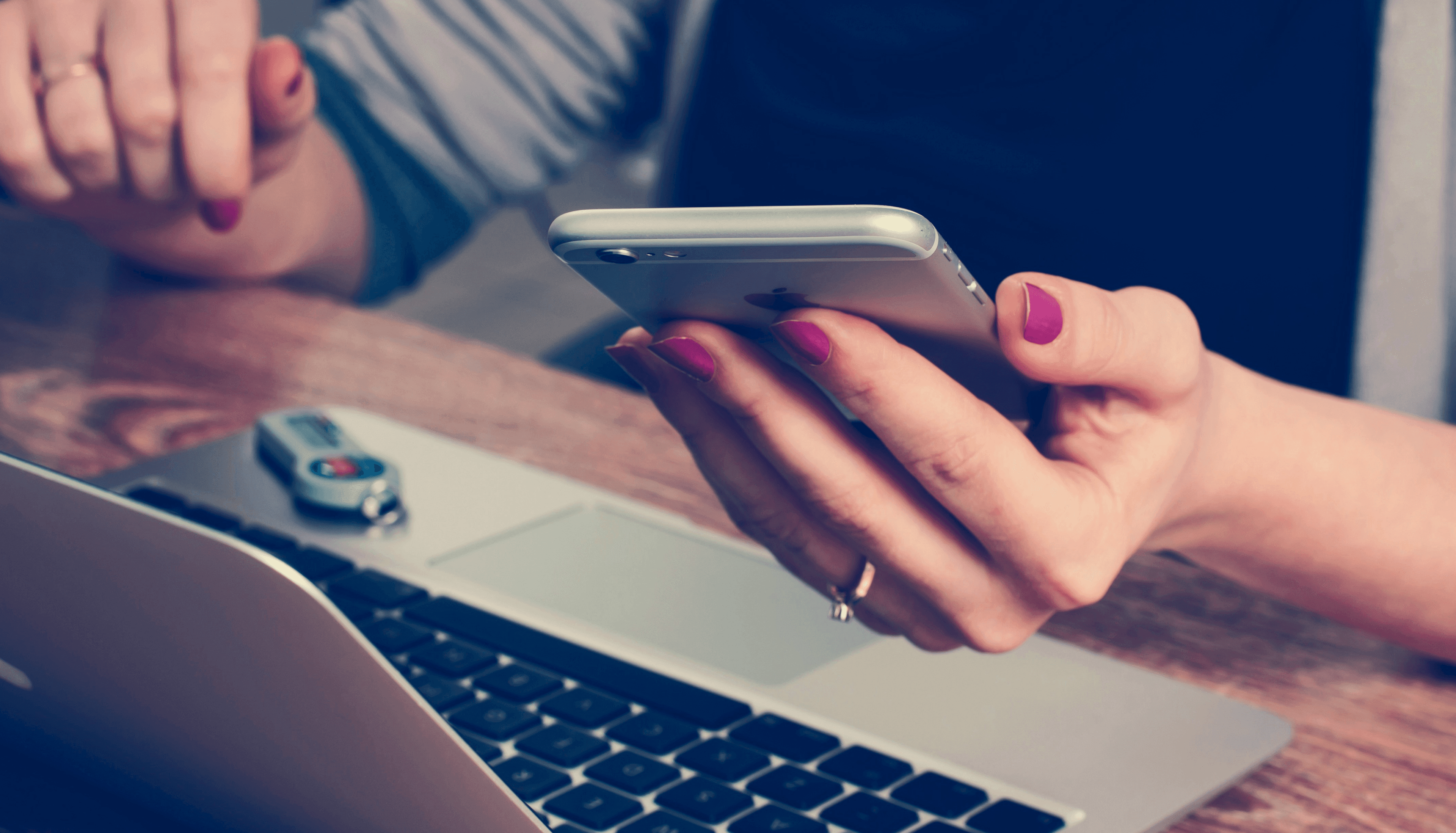 Female hand holding an iPhone above a laptop, nails painted pink, wearing golden rings.