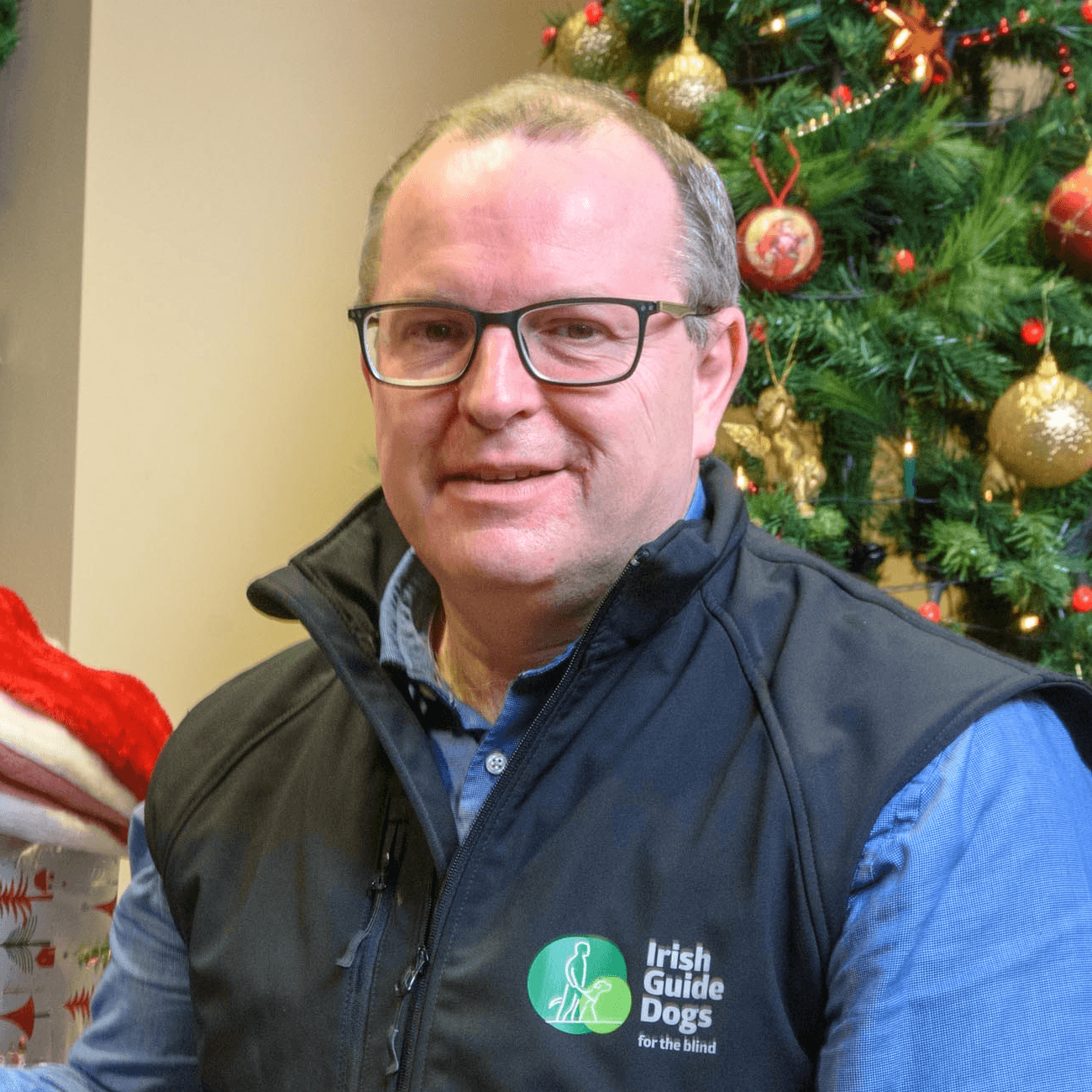 Headshot of Tim O'Mahony, CEO of the Irish Guide Dogs for the Blind, wearing a blue shirt and a black vest with the charity's logo on it.