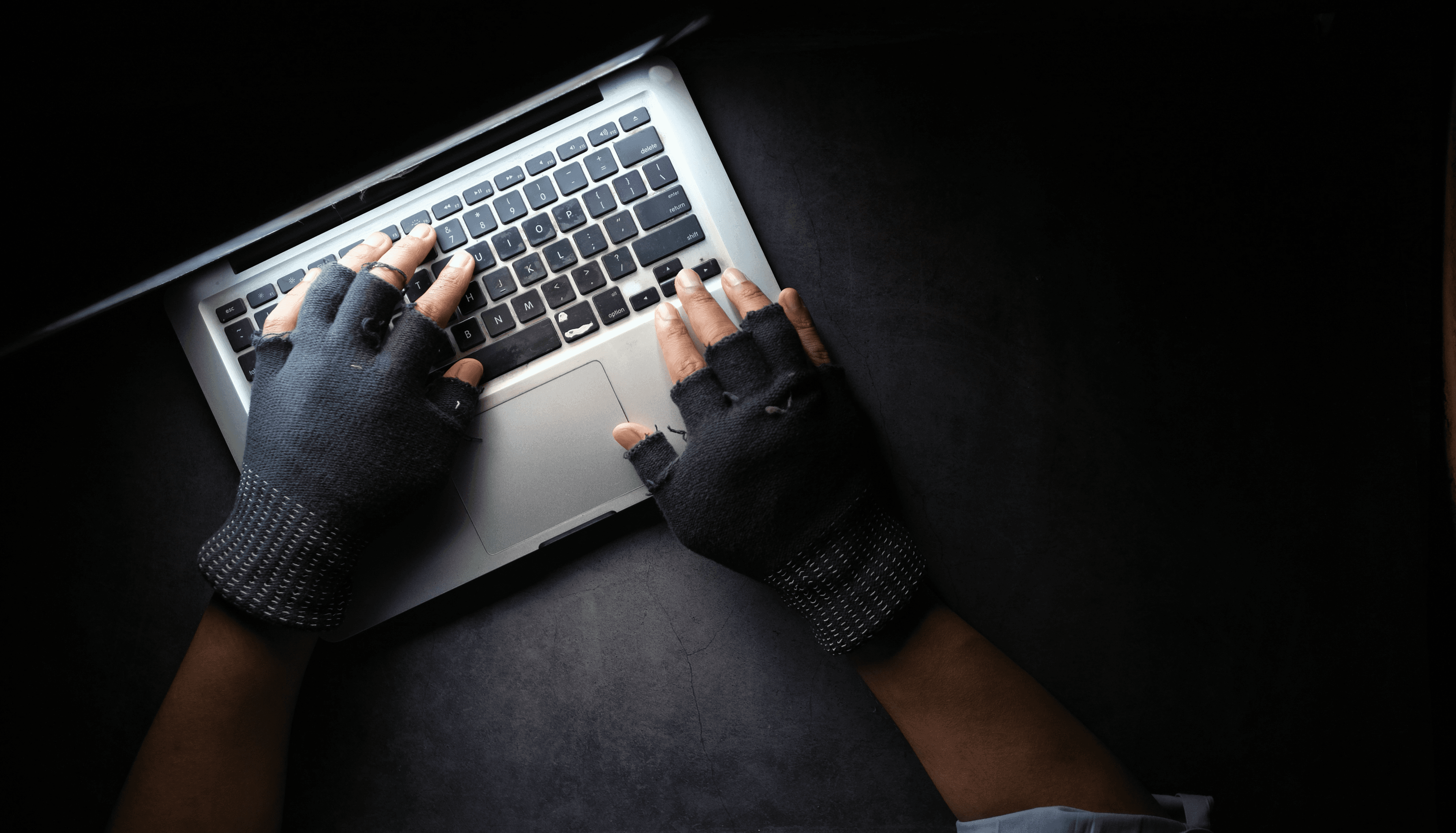 A pair of hands in fingerless gloves, suggesting criminal activity, typing on a laptop in a dimly lit room, with the laptop screen being the sole source of light.