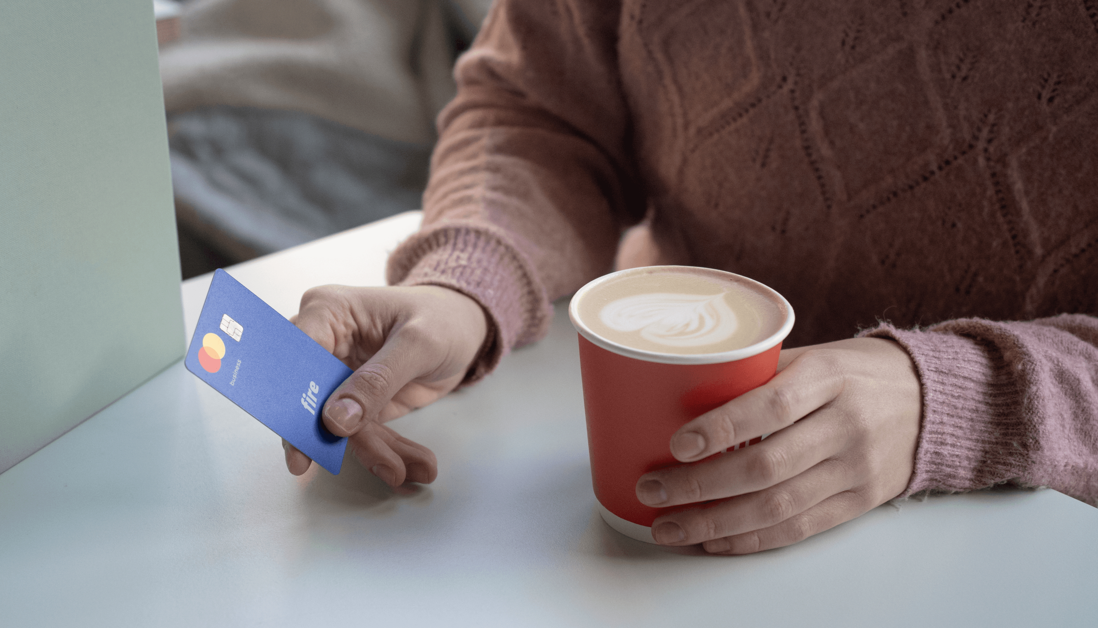 Close-up of a person holding a Fire business debit card in one hand and a paper coffee cup in the other, dressed in a pink jumper. The image suggests the act of tipping while purchasing a coffee.