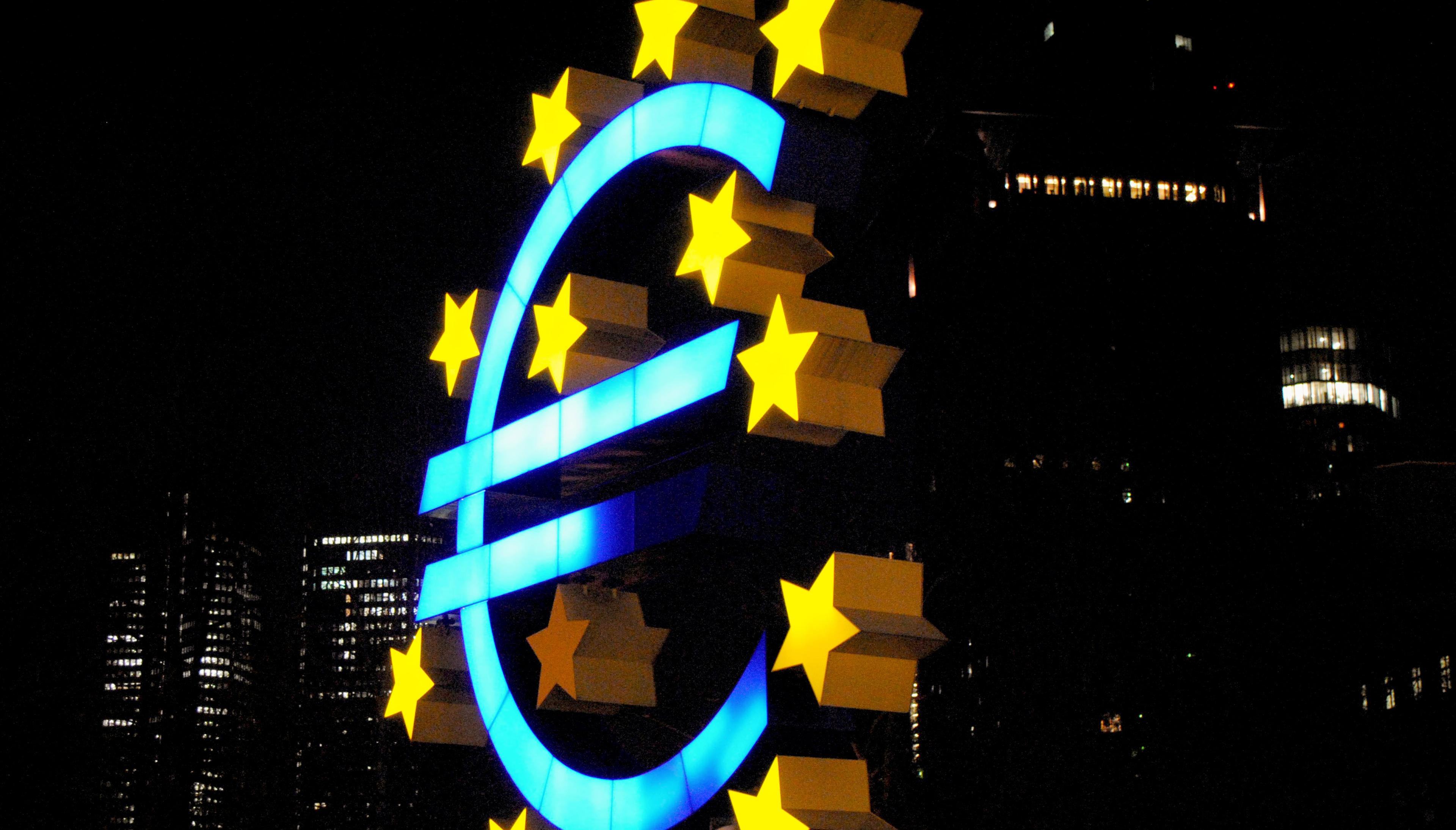 Statue of the euro currency symbol (€) with stars around it, set against a city skyline at night.