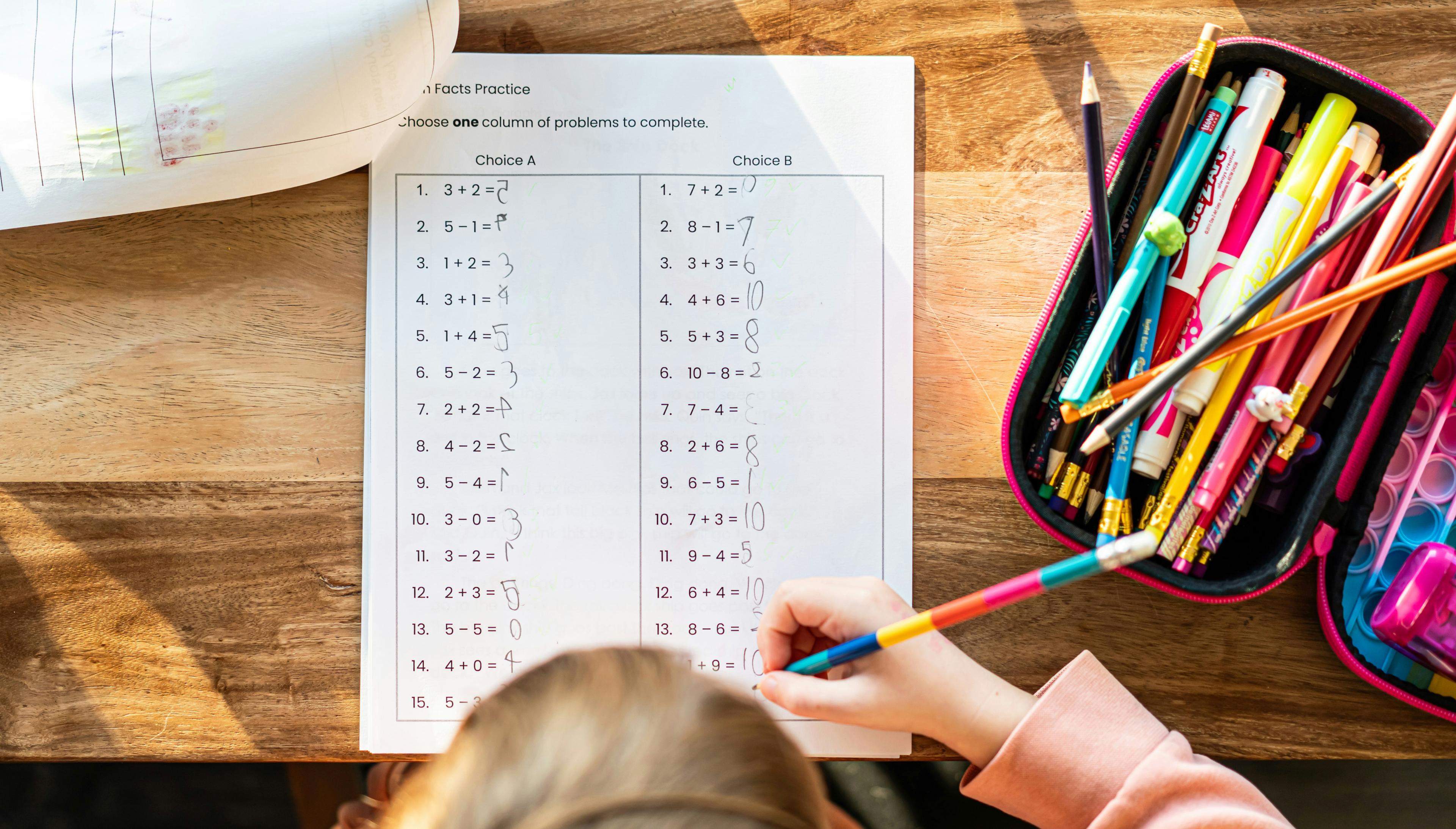 Child writing on a school worksheet on a wooden table, with a pencil case and other school supplies nearby.