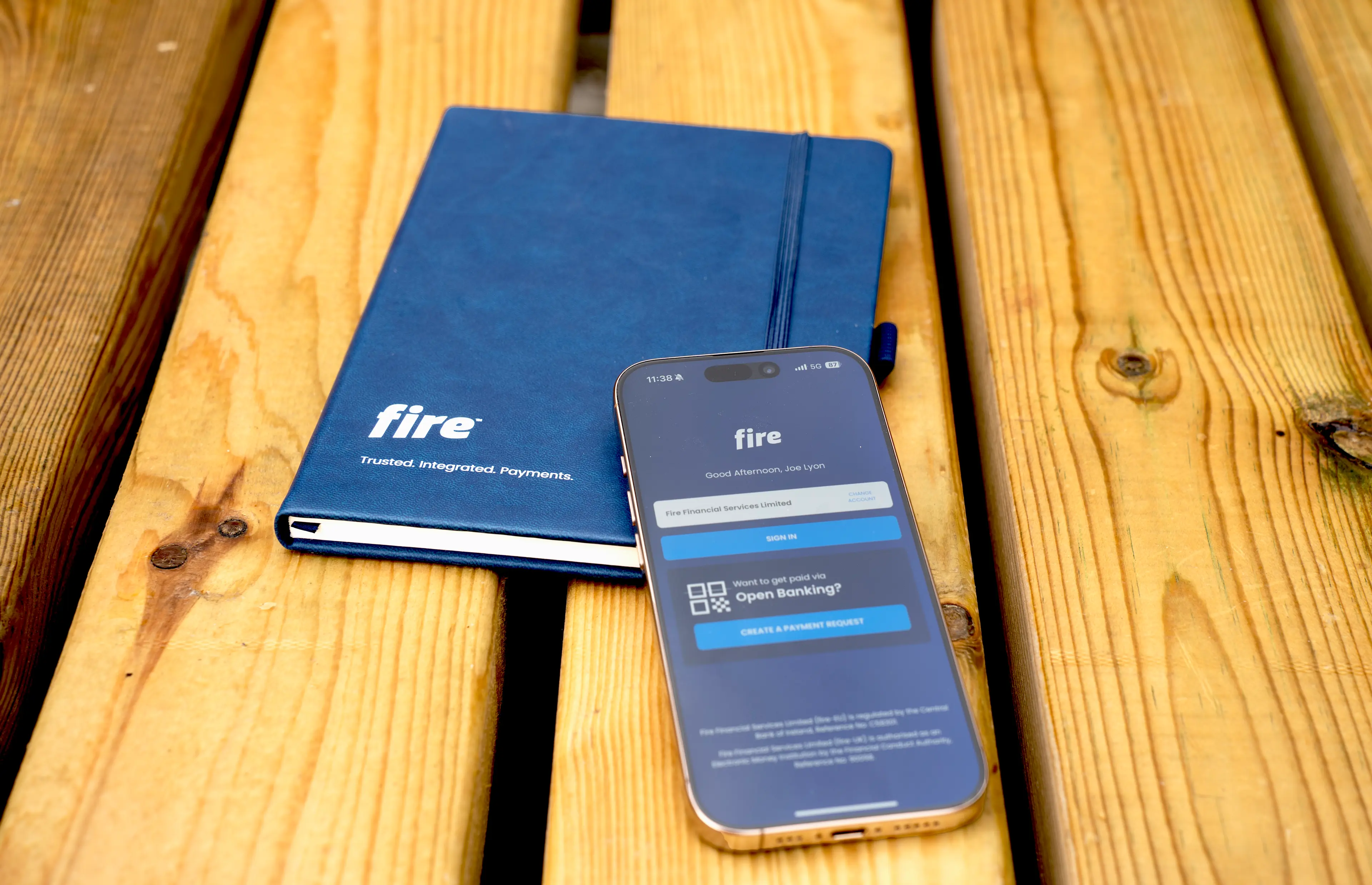 Phone displaying Fire’s login screen and an open banking payment request, placed over a notepad featuring Fire’s logo and the slogan “Trusted. Integrated. Payments.”